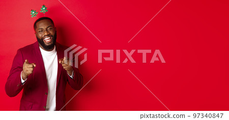 Merry Christmas. Handsome african american man in blazer and party headband, celebrating new year, pointing at camera and smiling, congratulating, red background 97340847