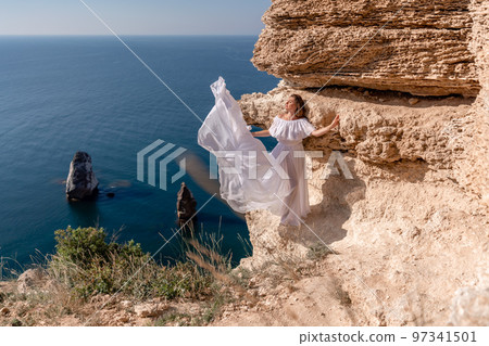 A beautiful young woman in a white light dress with long legs stands on the edge of a cliff above the sea waving a white long dress, against the background of the blue sky and the sea. 97341501