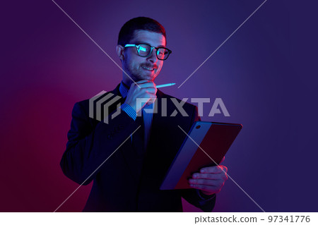 Young man, manager wearing business suit and eyeglasses using digital tablet on gradient blue purple background in neon light. Studying, work, job, finance 97341776