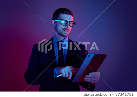 Young man, manager wearing business suit and eyeglasses using digital tablet on gradient blue purple background in neon light. Studying, work, job, finance Young man, manager wearing business suit and eyeglasses using digital tablet on gradient blue purple background in neon light. Studying, work, job, finance 97341791