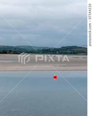 View of Clonakilty Bay at low tide. A small orange buoy is kept on a calm water surface. The sandy shore of the sea bay. The coastline on a day. 97344218