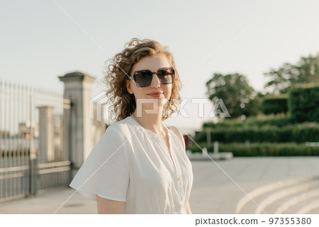 A woman with curly hair in sunglasses is posing in the park A woman with curly hair in sunglasses is posing in the park 97355380