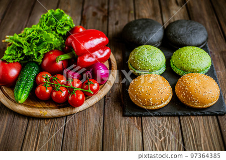 High angle view of variety of six hamburger buns with vegetables on the table. Black, green, yellow 97364385