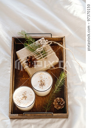 Christmas cozy morning in bed. Gift box and eggnog on wooden tray on white bedding. Christmas cozy morning in bed. Gift box and eggnog on wooden tray on white bedding. 97370150