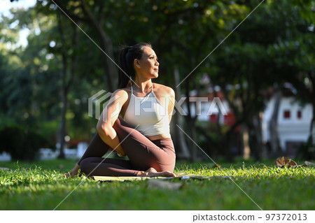 Millennial woman in sportswear sitting in Half lord of the fishes pose, practicing yoga on green park with sunlight 97372013