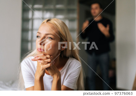 Close up face of unhappy tired woman sadly looking away sitting on bed on background of aggressive husband shouting on wife at home. Concept of family scandal, crisis, domestic violence, abuse. Close up face of unhappy tired woman sadly looking away sitting on bed on background of aggressive husband shouting on wife at home. Concept of family scandal, crisis, domestic violence, abuse. 97376063