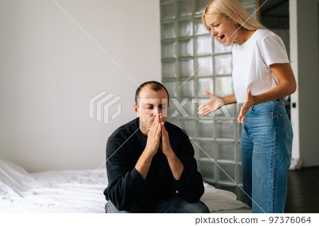 Portrait of tired depressed husband sitting on bed sadly looking away ignoring angry wife arguing blaming upset man of problems at home. Concept of family scandal, crisis, domestic violence, abuse. 97376064