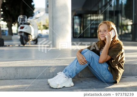 Teen girl sitting on street stairs on summer day and having mobile conversation on smartphone 97378239