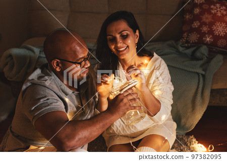 Happy interracial couple celebrating New Year at home together, sitting by Christmas tree Happy interracial couple celebrating New Year at home together, sitting by Christmas tree 97382095