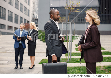 Close up on caucasian woman wearing black jacket and talking with black man. Confident businesswoman and man standing in front of modern office building. Banking and financial market concept. Close up on caucasian woman wearing black jacket and talking with black man. Confident businesswoman and man standing in front of modern office building. Banking and financial market concept. 97384734