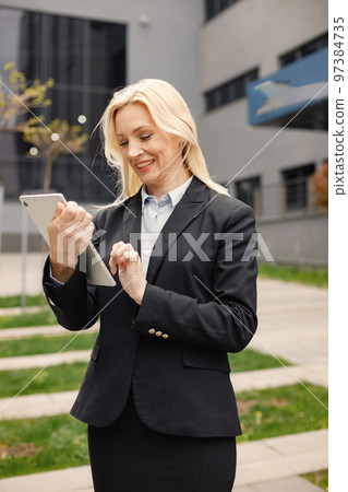 Close up on caucasian woman wearing black jacket and using a tablet. Confident businesswoman standing in front of modern office building. Banking and financial market concept. Close up on caucasian woman wearing black jacket and using a tablet. Confident businesswoman standing in front of modern office building. Banking and financial market concept. 97384735