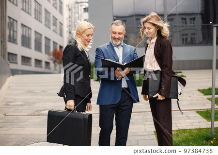 Confident businesspersons standing in front of modern office building. Man and women talking and holding documents. Banking and financial market concept. Confident businesspersons standing in front of modern office building. Man and women talking and holding documents. Banking and financial market concept. 97384738