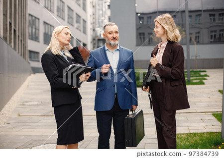 Confident businesspersons standing in front of modern office building. Man and women talking and holding documents. Banking and financial market concept. 97384739