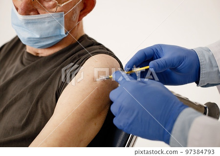 Cropped photo of senior gentleman. Man wearing khaki t-shirt and face mask. Doctor's hands in blue gloves doing an injection. Cropped photo of senior gentleman. Man wearing khaki t-shirt and face mask. Doctor's hands in blue gloves doing an injection. 97384793