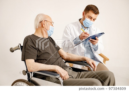 Senior gentleman on a wheelchair isolated on a white background. Man wearing khaki t-shirt and doctor wearing white medical gown and face mask. Doctor talking with man and fill in an informed consent. 97384809