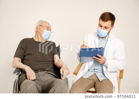 Senior gentleman on a wheelchair isolated on a white background. Man wearing khaki t-shirt and doctor wearing white medical gown and face mask. Doctor talking with man and fill in an informed consent. 97384811