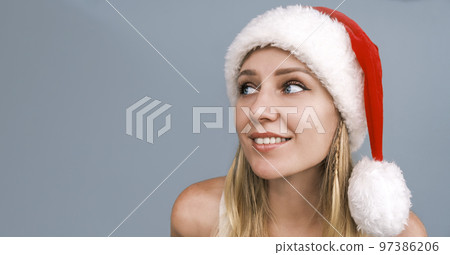 Dreamy smiling Santa girl wearing red hat looking side up. Portrait of beautiful young blue-eyed blonde woman in Santa's hat with bare shoulders. Christmas portrait. Christmas holidays or vacation. Dreamy smiling Santa girl wearing red hat looking side up. Portrait of beautiful young blue-eyed blonde woman in Santa's hat with bare shoulders. Christmas portrait. Christmas holidays or vacation. 97386206