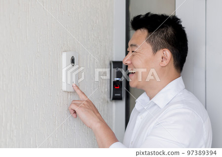 Close-up photo. A young Asian man rings an electronic doorbell on the wall of a house with his finger. He opens the door to his home. 97389394