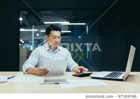 Portrait of a young male Asian accountant. He sits at the desk in the office, works with documents, accounts on a calculator and a laptop. Portrait of a young male Asian accountant. He sits at the desk in the office, works with documents, accounts on a calculator and a laptop. 97389396