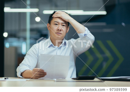 Shocked male Asian student holds a document in his hands. The worried person holds his head. Sitting at the desk in the office, received a letter, exam results, information about studies, bill. 97389399