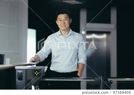 Young handsome Asian male worker enters office through electronic turnstile, automatic security system, gate. Encloses his electronic access card. He looks at the camera, smiles. 97389408