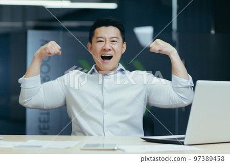 Portrait of a young happy Asian man at the desk in the office celebrating success, victory. He looks at the camera, shows a gesture of strength with his hands. Portrait of a young happy Asian man at the desk in the office celebrating success, victory. He looks at the camera, shows a gesture of strength with his hands. 97389458