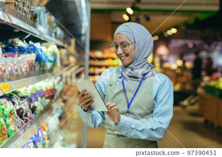 woman Seller in super market in hijab with tablet checking products using pocket computer, Muslim woman near shelves with products and goods. 97390451