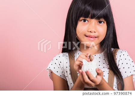 Asian little kid 10 years old holding piggy bank and looking at camera at studio shot isolated on pink background, Happy child girl lifestyle smiling with is full piggybank, Personal money savings 97390874