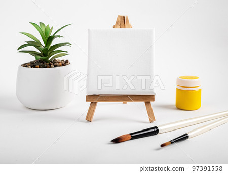 Clean blank canvas mockup for painting, drawing on easel, paint brushes, potted plant and yellow gouache. High quality photo Clean blank canvas mockup for painting, drawing on easel, paint brushes, potted plant and yellow gouache. High quality photo 97391558