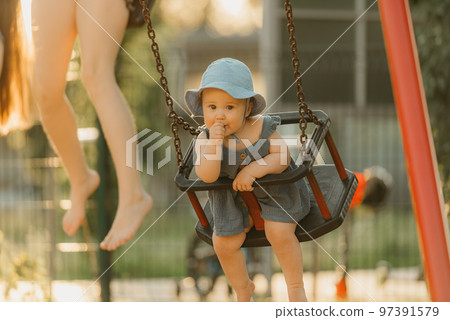 Toddler in a dress is sucking her thumb on a swing on the warm summer evening 97391579