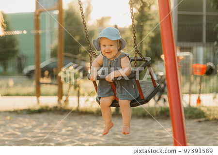 Toddler baby girl in a dress is happy on a swing on the warm summer evening 97391589
