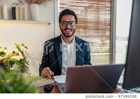 Smiling indian businessman in suit working on project from home office and looking at camera Smiling indian businessman in suit working on project from home office and looking at camera 97399398