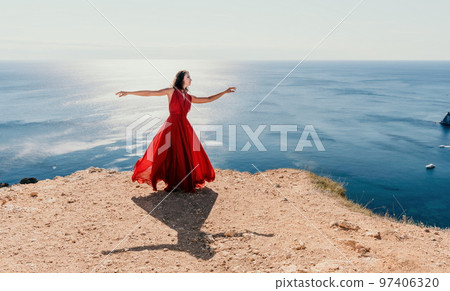 Woman in red dress on sea. Side view a Young beautiful sensual woman in a red long dress posing on a rock high above the sea on sunset. Girl on the nature on blue sky background. Fashion photo. Woman in red dress on sea. Side view a Young beautiful sensual woman in a red long dress posing on a rock high above the sea on sunset. Girl on the nature on blue sky background. Fashion photo. 97406320