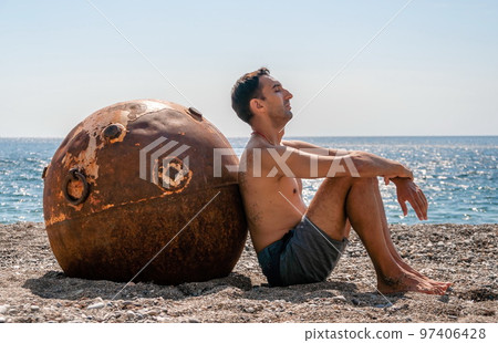 Man doing yoga and meditation outdoors near to old rusty floating marine mine on the beach with rocky shore and sea background. Healthy lifestyle, pollution, nature protection, war and peace concept. 97406428