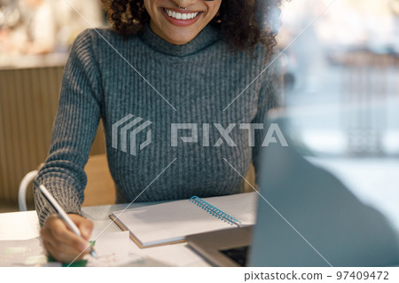 Smiling woman freelancer working laptop and making notes while sitting near windows in cozy cafe Smiling woman freelancer working laptop and making notes while sitting near windows in cozy cafe 97409472