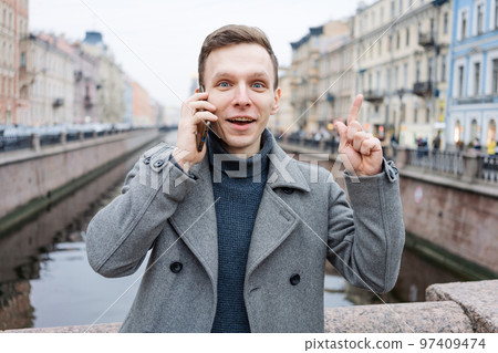 Emotional young man using mobile phone, wearing an elegant gray coat, stands 97409474
