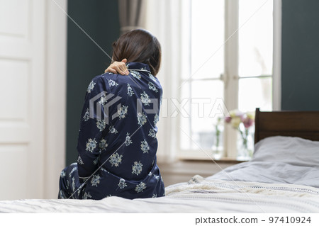 A middle-aged woman in pajamas sitting on the bed and holding her shoulders Back view A middle-aged woman in pajamas sitting on the bed and holding her shoulders Back view 97410924