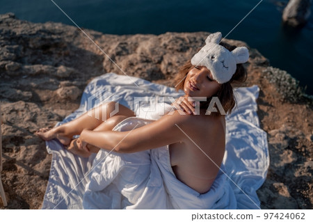 Young woman in sleep mask posing on the seashore. She is wrapped in a white blanket. The coast of the sea at a height, around the sea and rocks, dawn. 97424062