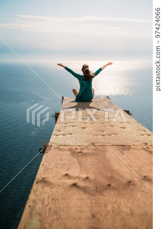 A girl sits on a wooden springboard for jumping with a rope. In a dark green dress with her hands in the air. A girl sits on a wooden springboard for jumping with a rope. In a dark green dress with her hands in the air. 97424066