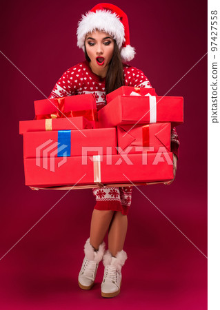 Amazed beautiful woman holding many presents on red background. Young woman in santa hat looking at boxes Amazed beautiful woman holding many presents on red background. Young woman in santa hat looking at boxes 97427558