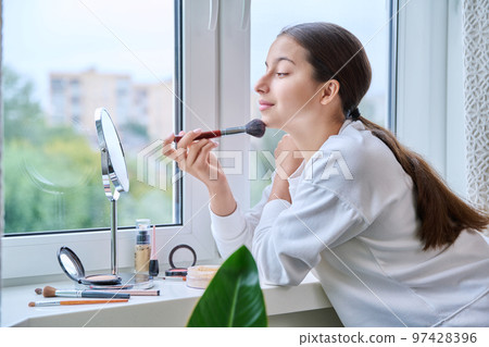 Young teenage girl with cosmetics mirror doing natural makeup, at home Young teenage girl with cosmetics mirror doing natural makeup, at home 97428396