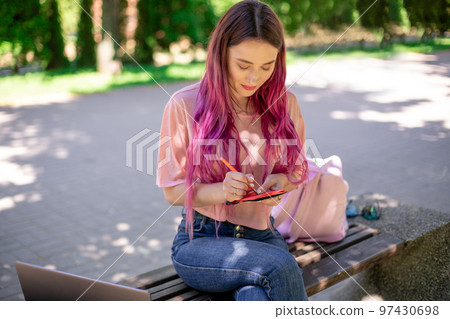 Woman writing in a notebook sitting on a wooden bench in the park. Girl working outdoors on portable computer, copy space. Woman writing in a notebook sitting on a wooden bench in the park. Girl working outdoors on portable computer, copy space. 97430698