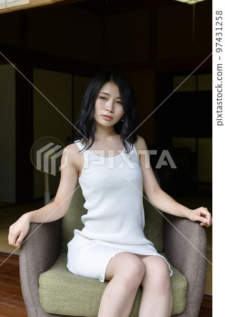A young woman in a white dress sitting on a sofa on the porch of an old private house 97431258