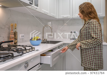 Cute young housewife pulls out the cabinet drawer at kitchen Cute young housewife pulls out the cabinet drawer at kitchen 97431764