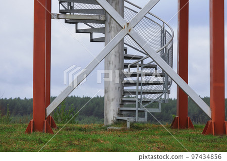 Sirveta Observation Tower bottom with stairs on cloudy sky background during the dry autumn season with green grass and row of forest trees on the background 97434856