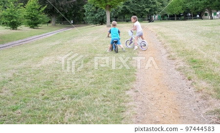 Sport activity for children. Small kids playing bicycle and scooter riding on path in park in summer drought dry grass. Little Brother and Sister Siblings playing together. Child development concept Sport activity for children. Small kids playing bicycle and scooter riding on path in park in summer drought dry grass. Little Brother and Sister Siblings playing together. Child development concept 97444583