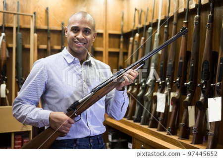 Happy hispanic man standing with hunting rifle at gun shop 97445652