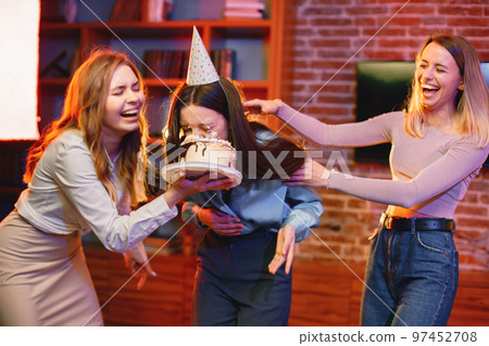 Young woman dips face in white cake with cream. Three girls having fun together. Birthday face cake smashing tradition concept. Young woman dips face in white cake with cream. Three girls having fun together. Birthday face cake smashing tradition concept. 97452708