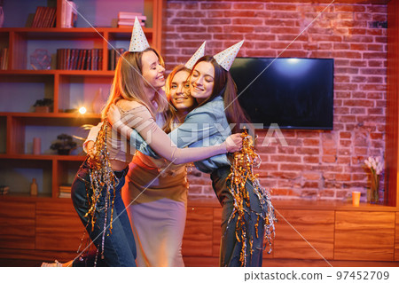 Young friends standing in indoors in evening party red light. Girls having fun under a confetti. Everybody wearing party cones. Young friends standing in indoors in evening party red light. Girls having fun under a confetti. Everybody wearing party cones. 97452709