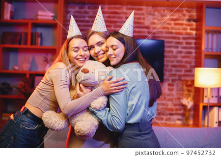 Young friends standing in indoors in evening party red light. Everybody wearing party cones. Girl holding a bear toy gift. Young friends standing in indoors in evening party red light. Everybody wearing party cones. Girl holding a bear toy gift. 97452732